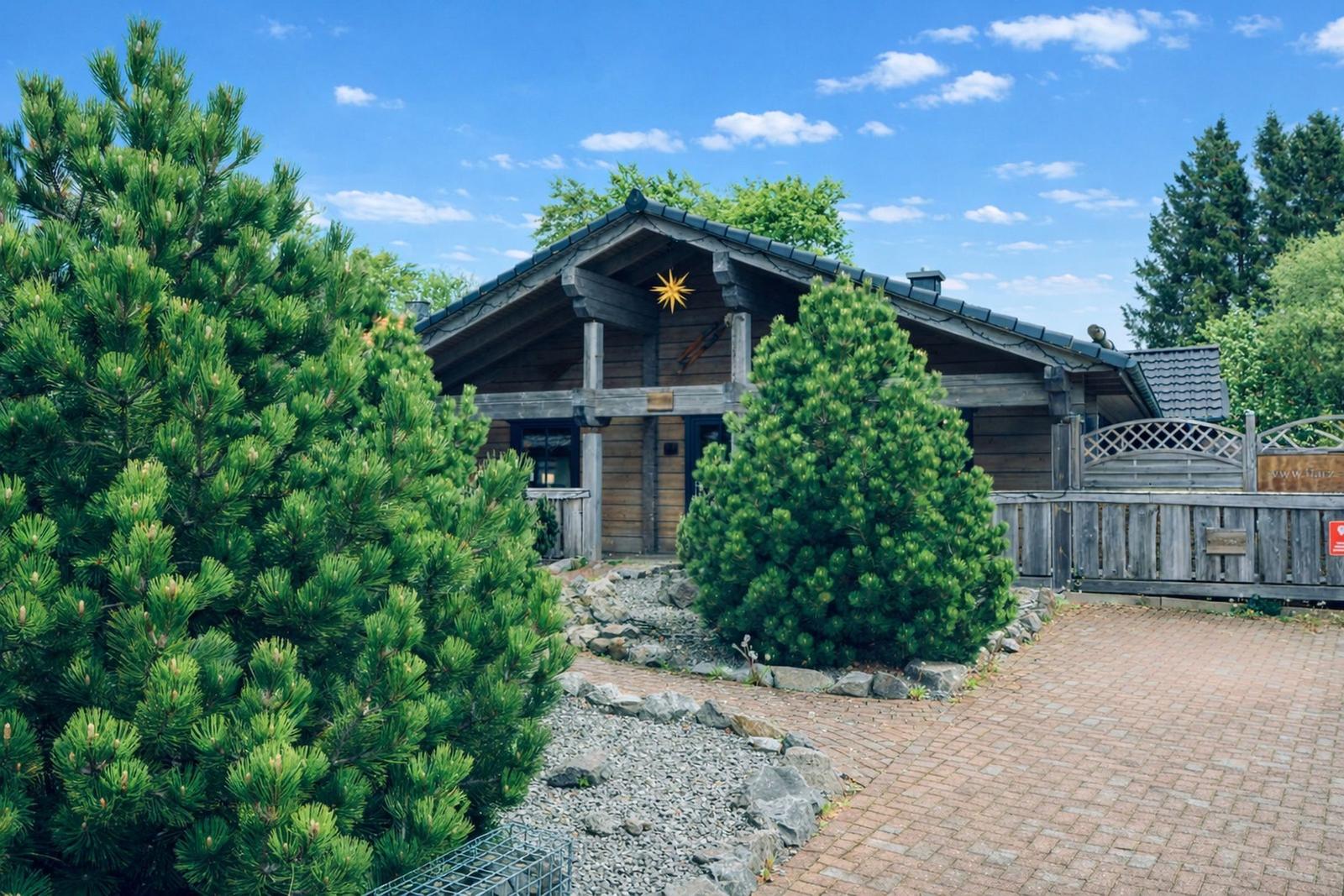 Holzhaus mit Steinweg und Nadelbäumen unter blauem Himmel.