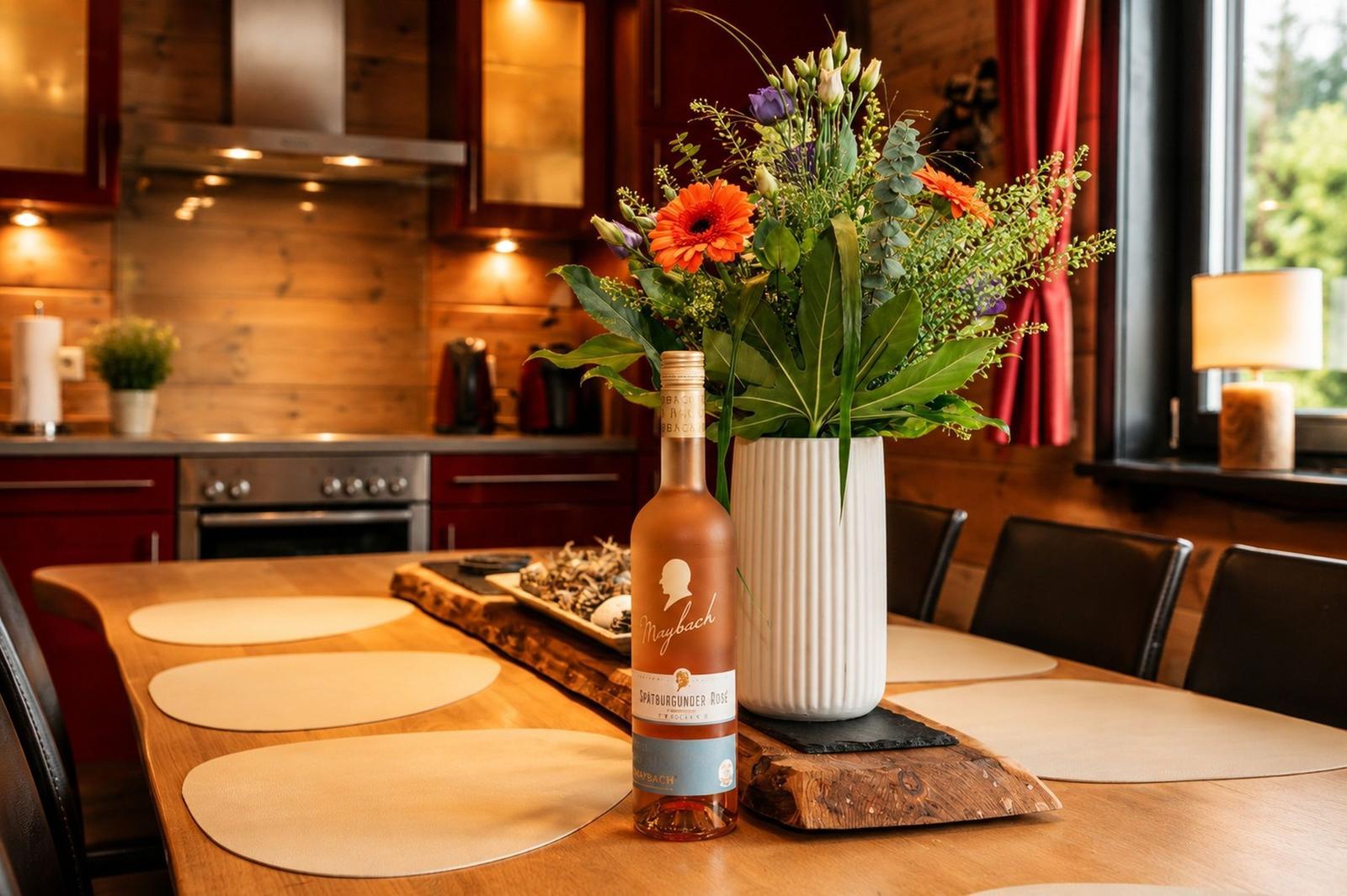 Esstisch mit Blumen und Weinflasche in rustikaler Küche mit Holzwänden und roten Schränken.