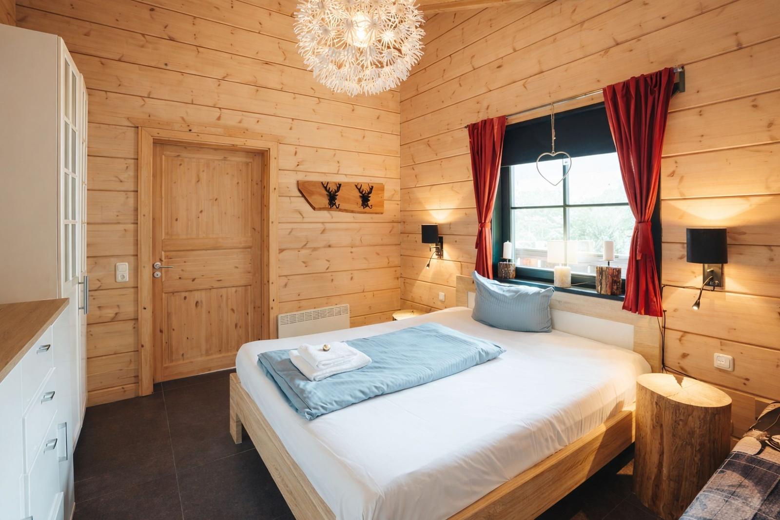 Schlafzimmer mit Holzwand, Bett, Fenster mit roten Vorhängen und Deckenleuchte.