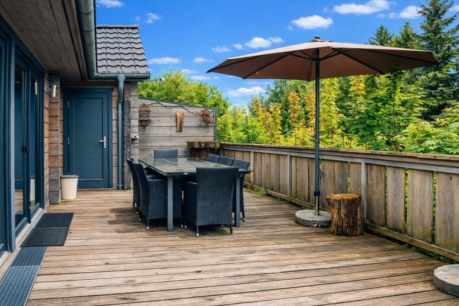 Terrasse mit Holzboden, Tisch, Stühlen und Sonnenschutz. Hinterhof mit Holzzaun und Bäumen.
