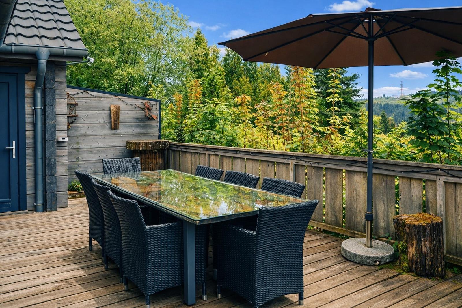 Terrasse mit Tisch, Stühlen und Sonnenschirm. Holzgeländer und grüne Vegetation im Hintergrund.