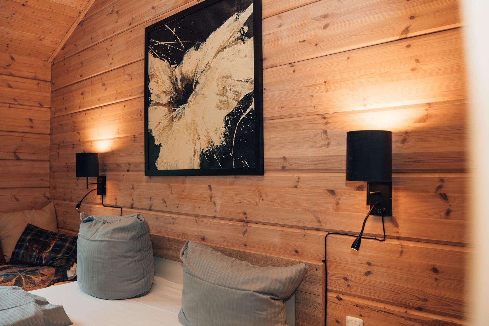 Schlafzimmer mit Holzwand, Bett, Kissen und Wandbild. Zwei Wandlampen beleuchten den Raum.