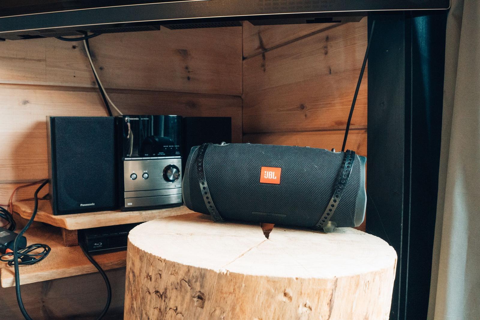 Ein JBL-Lautsprecher steht auf einem Baumstamm. Hinter ihm befindet sich ein Panasonic-Audio-Gerät.