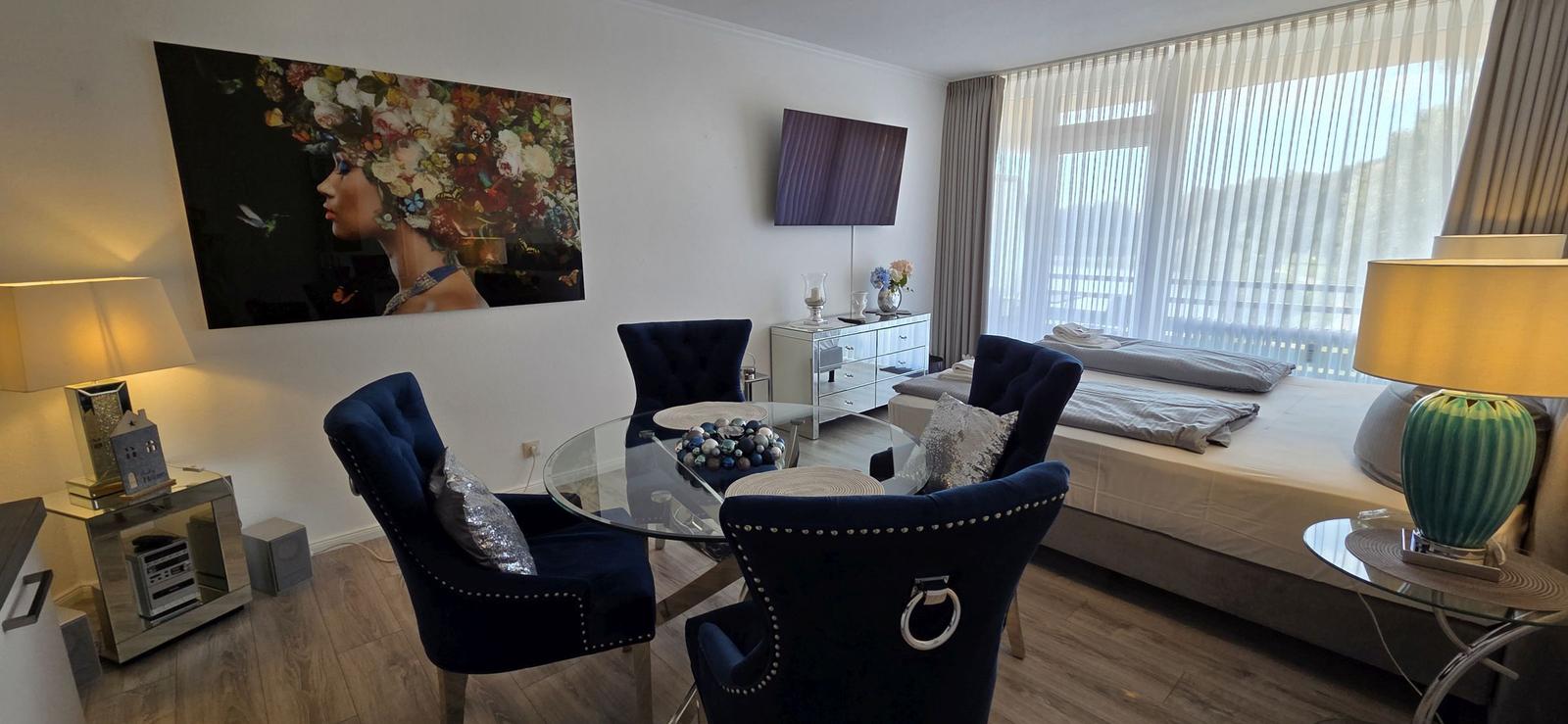 Schlafzimmer mit Schlafcouch, Glas-Tisch und blauen Stühlen. Große Fenster mit Jalousien.