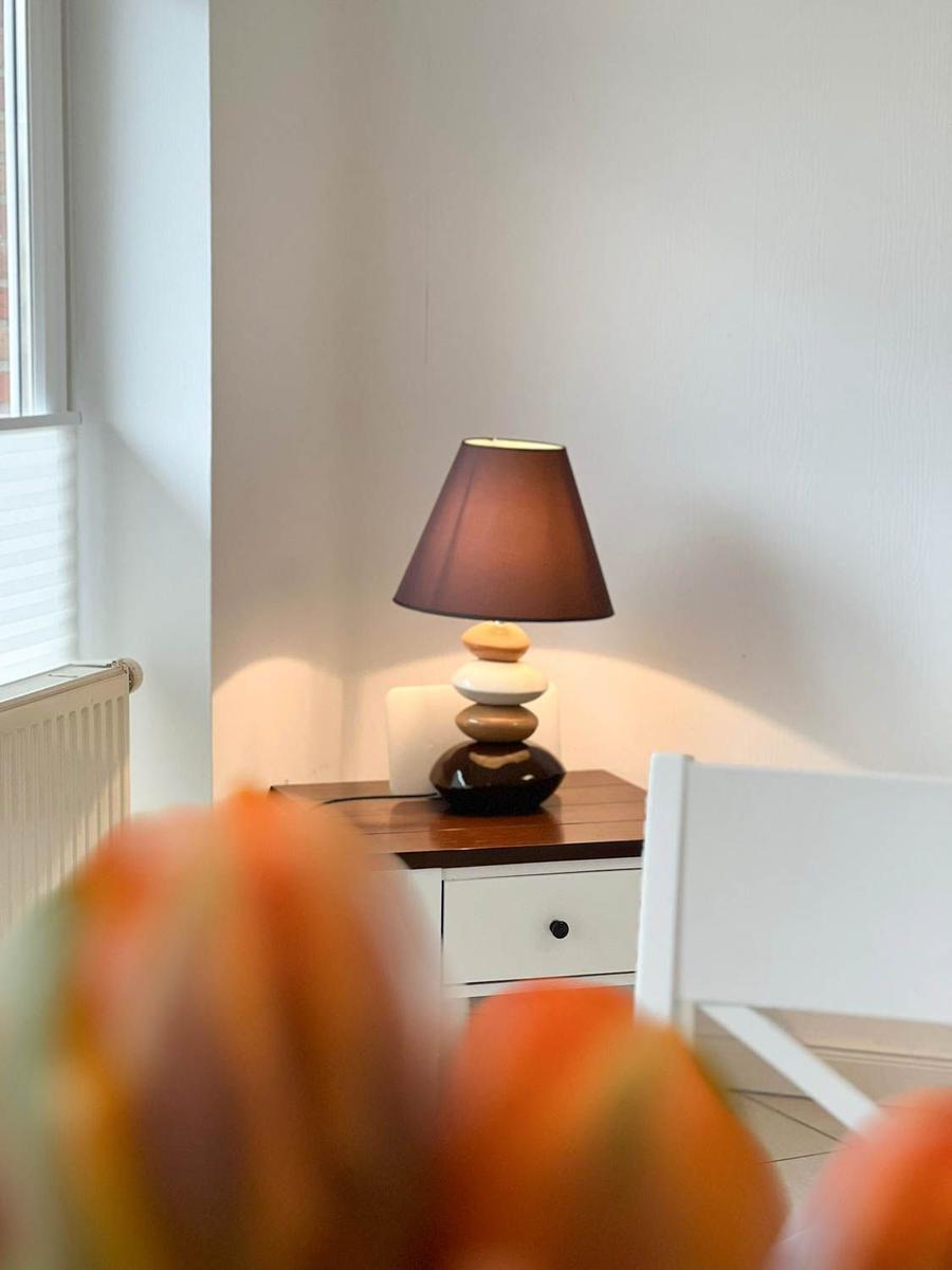 Tischlampe auf Nachttisch neben Fenster mit Heizkörper und Stuhl im Vordergrund.