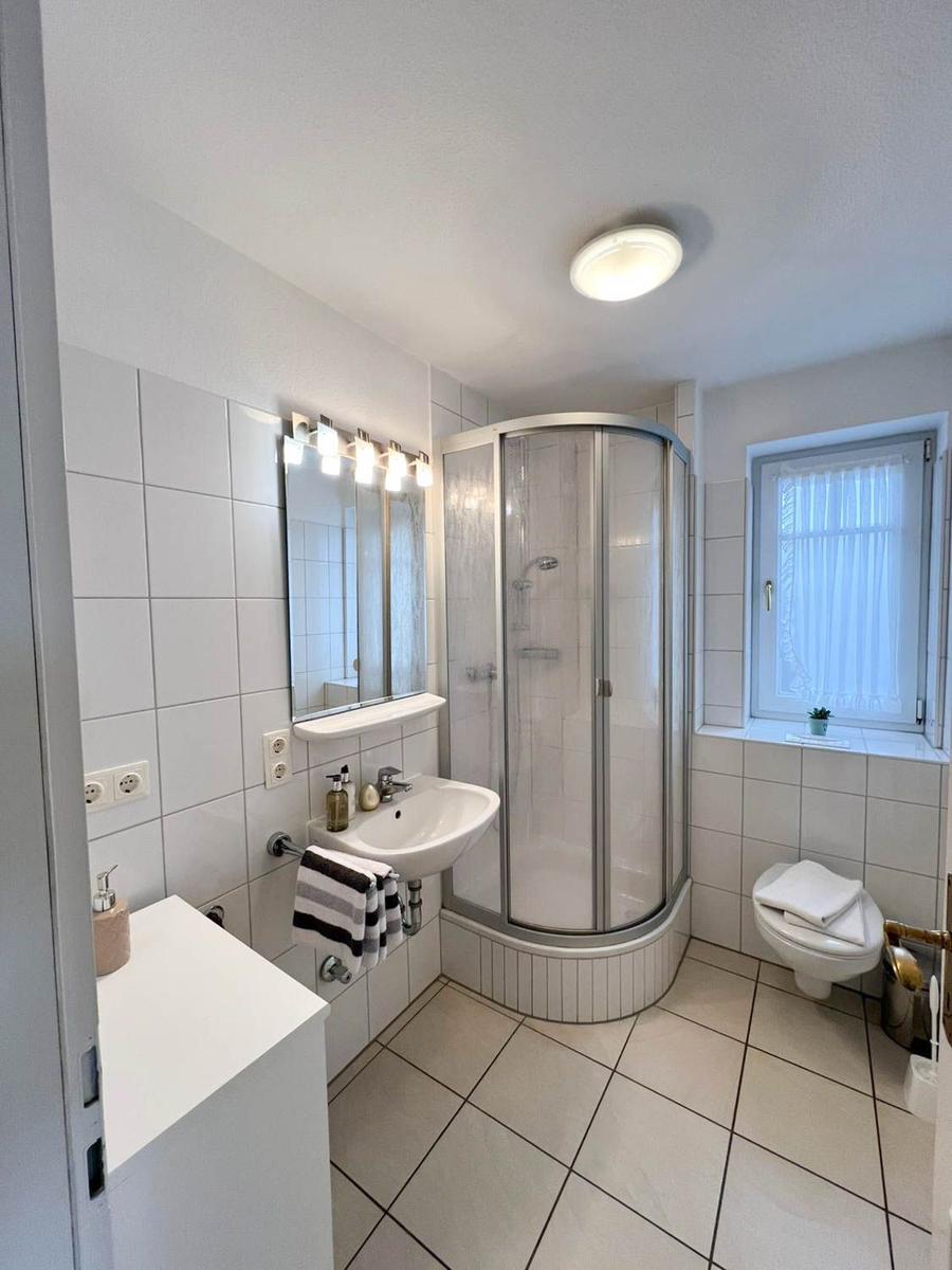 Badezimmer mit Dusche, Waschbecken und Toilette. Fenster mit Vorhang.