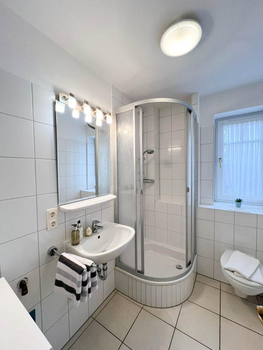 Badezimmer mit Dusche, Waschbecken und Toilette. Fenster mit Vorhang.