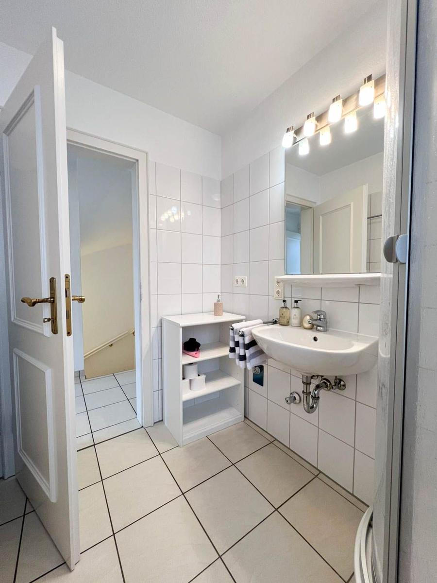 Badezimmer mit Waschbecken, Spiegel und Regal. Tür führt in einen Flur.