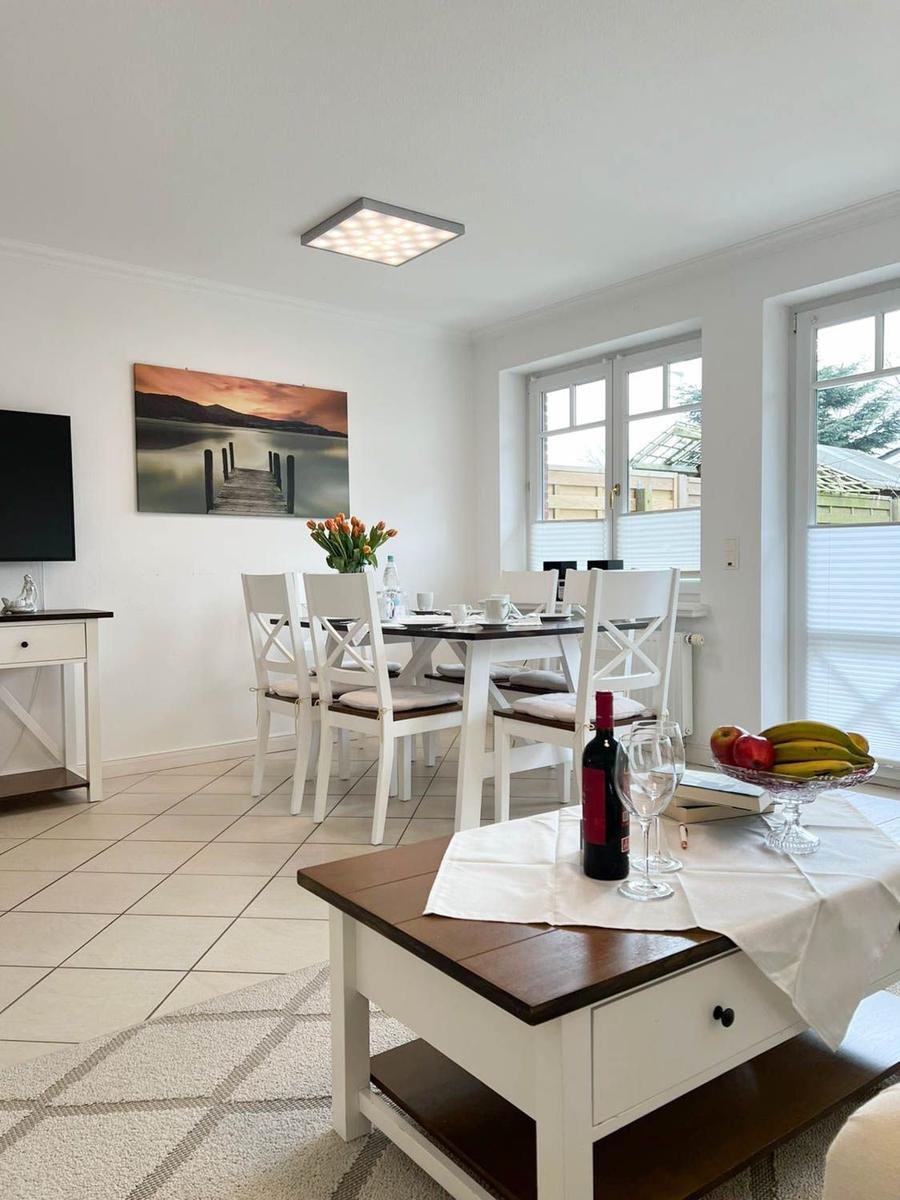 Esszimmer mit Tisch, Stühlen und Fenstern. Auf Tisch steht Wein und Obst.