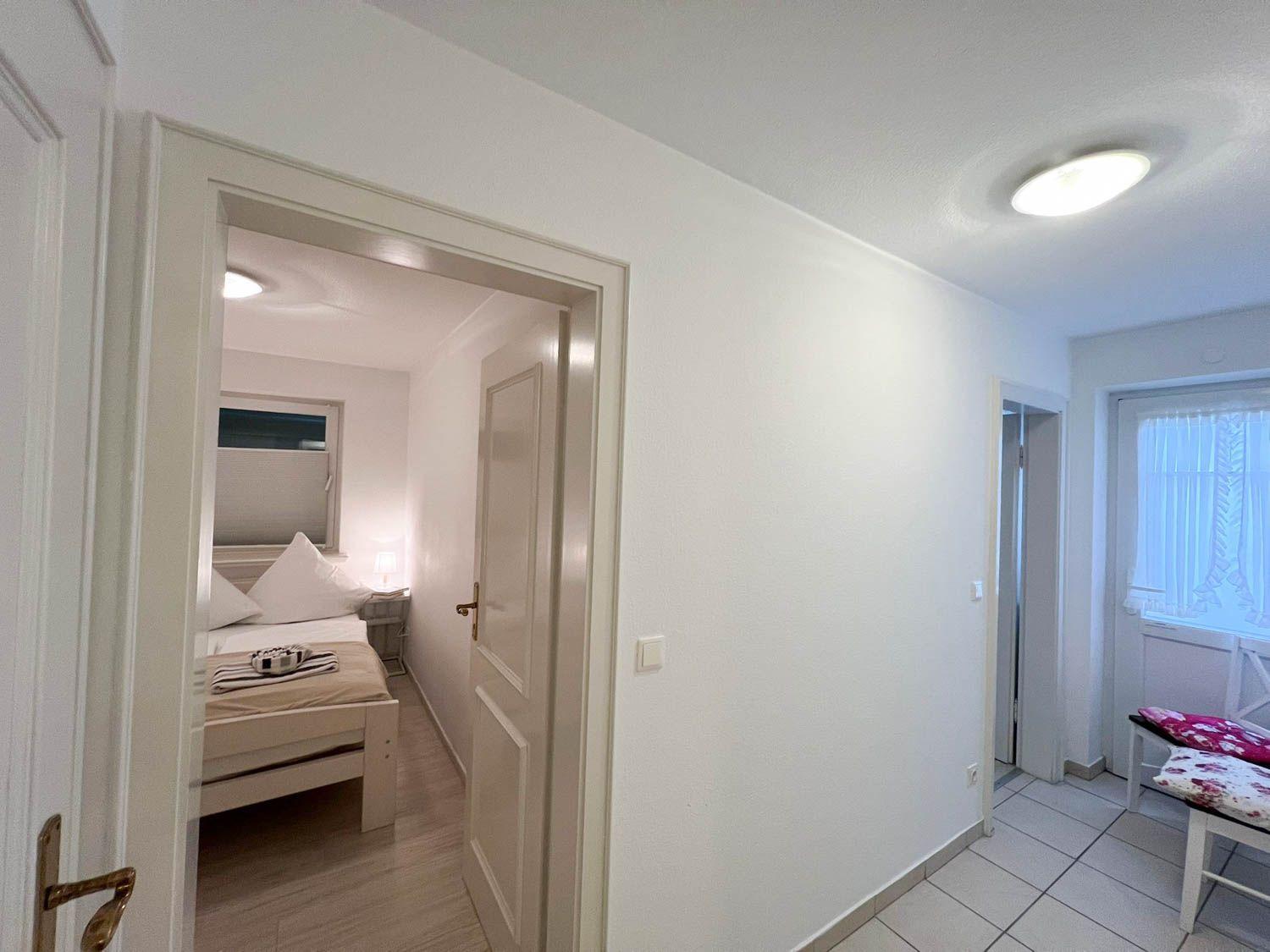 Schlafzimmer mit Bett und Nachttisch. Flur mit Tür und Fenster. Bodenfliesen und weiße Wände.