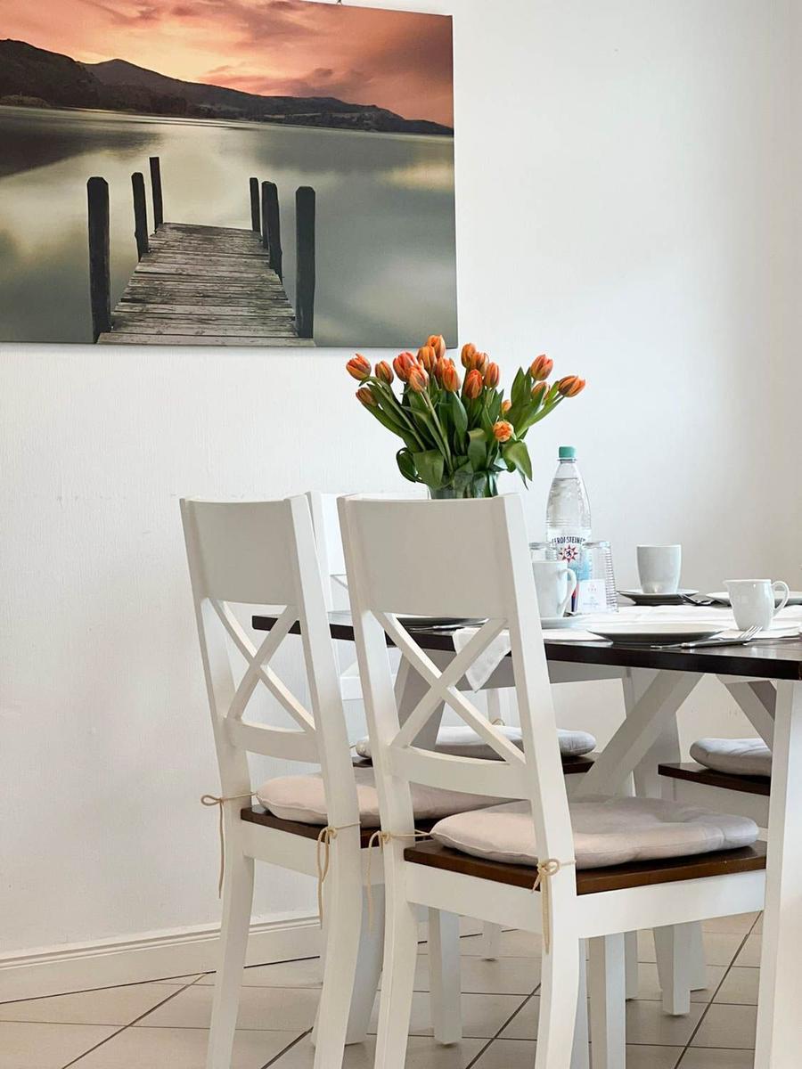 Esstisch mit weißen Stühlen und Tulpenblumen. Wandbild zeigt einen Steg über Wasser.
