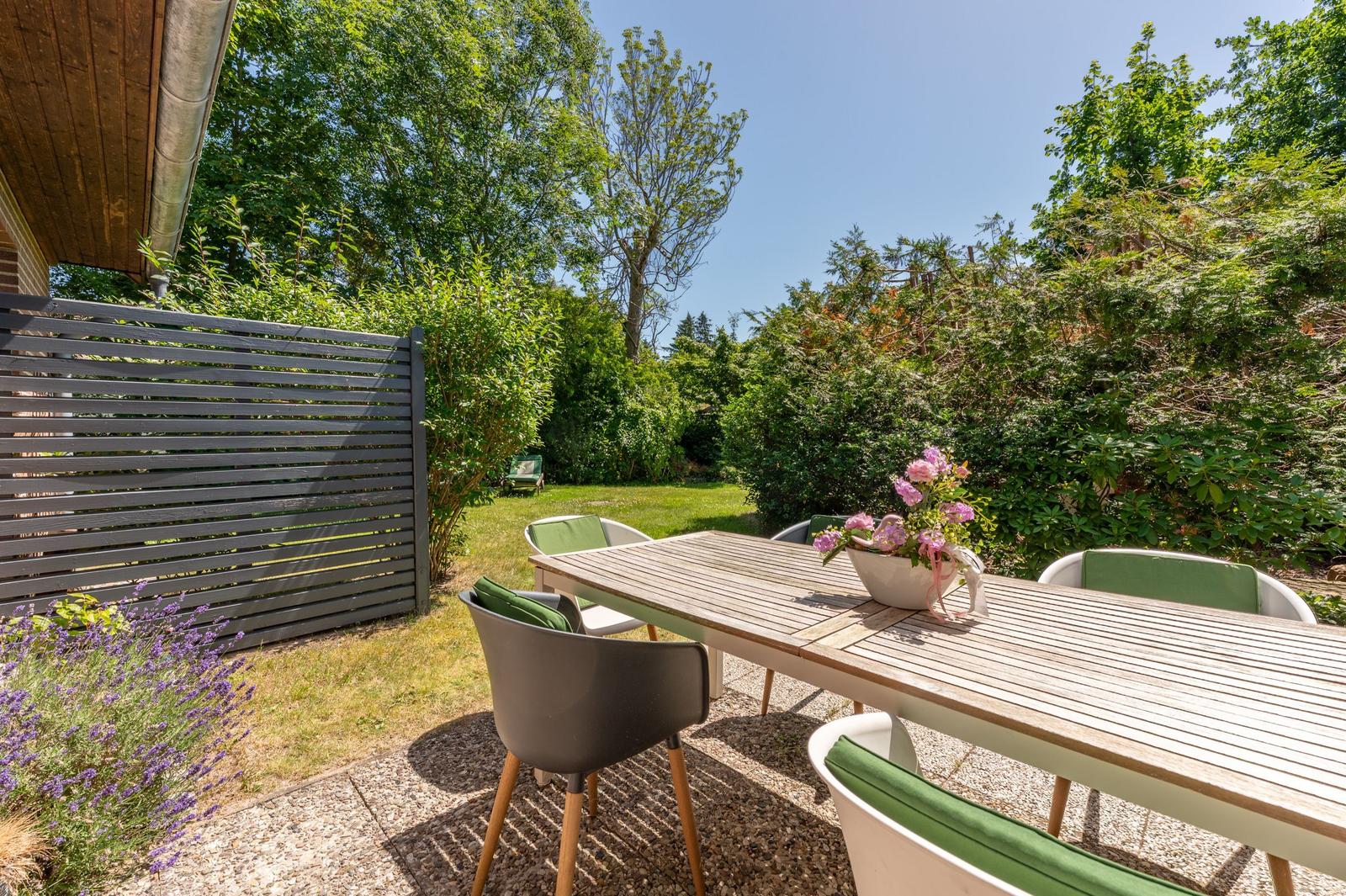 Hinterhof mit Holztisch, Stühlen und Blumen. Grünflächen und Holzgeländer im Hintergrund.
