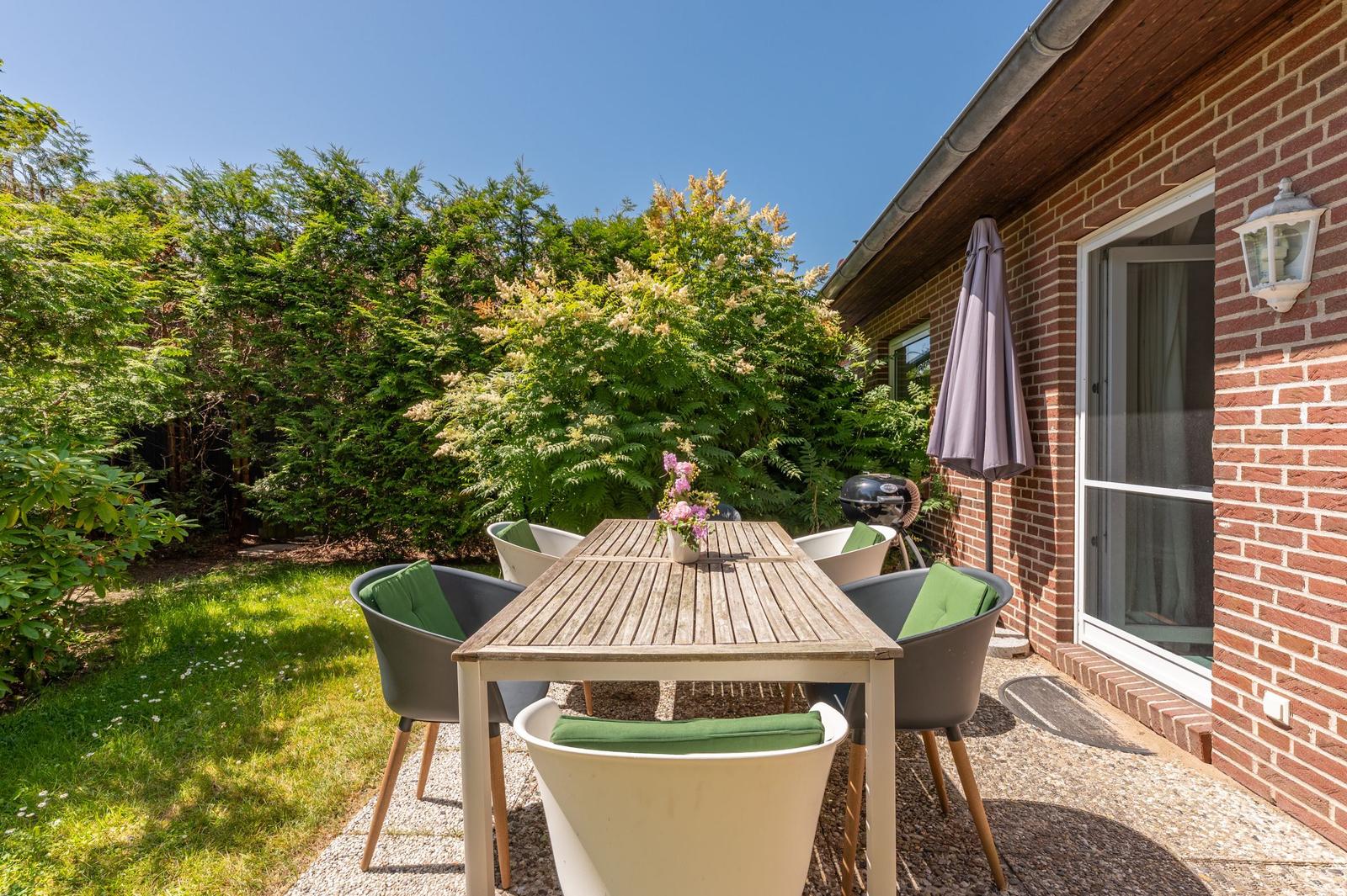 Terrasse mit Tisch, Stühlen und Grill. Hintergrund: grüne Hecke und Hauswand.