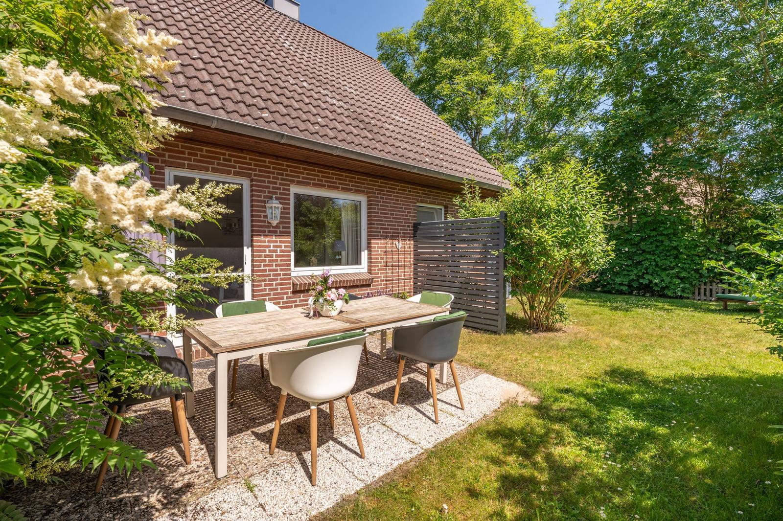 Hinterhof mit Tisch und Stühlen, Grünflächen und Blumen. Haus mit Ziegeldach und Fenstern.