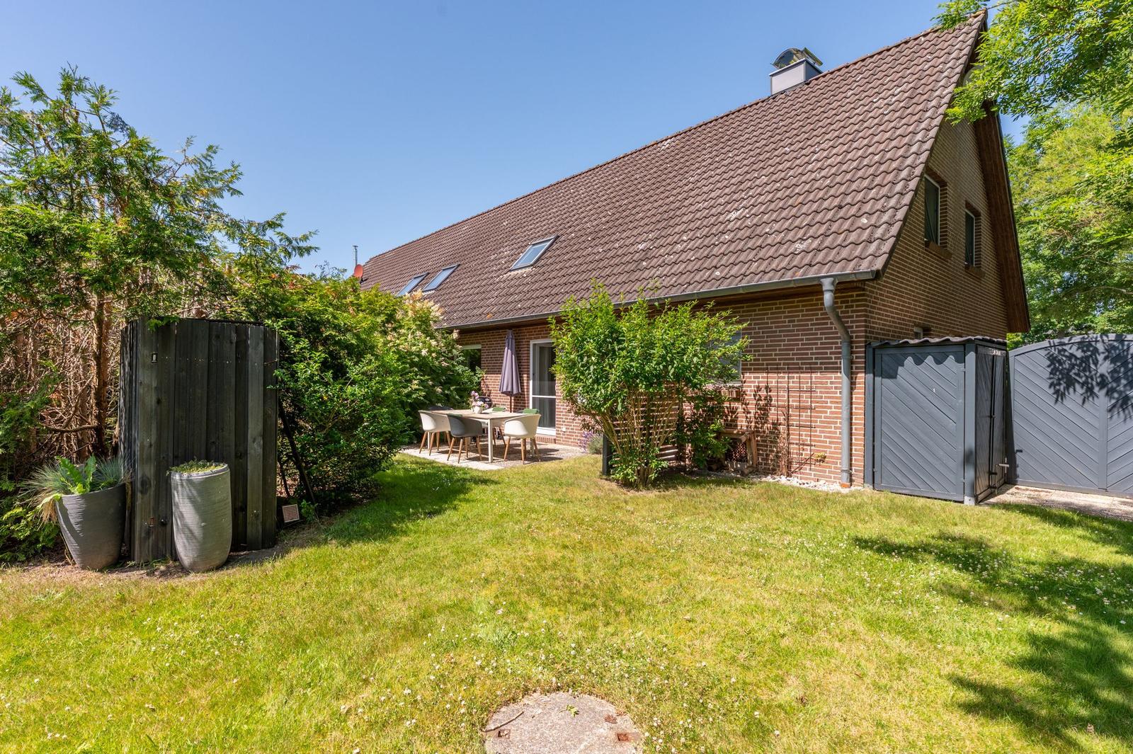 Haus mit Garten, Terrasse und Tisch mit Stühlen. Dach mit Dachfenstern und Schornstein.