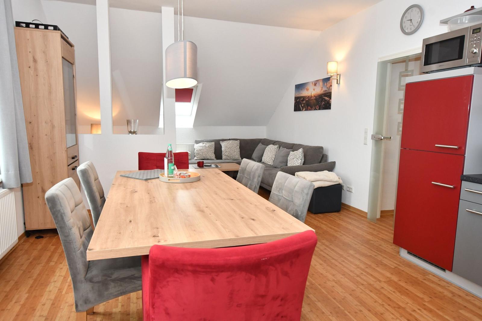Essbereich mit Holztisch und Stühlen. Im Hintergrund befindet sich ein Wohnbereich mit Sofa und Kühlschrank.