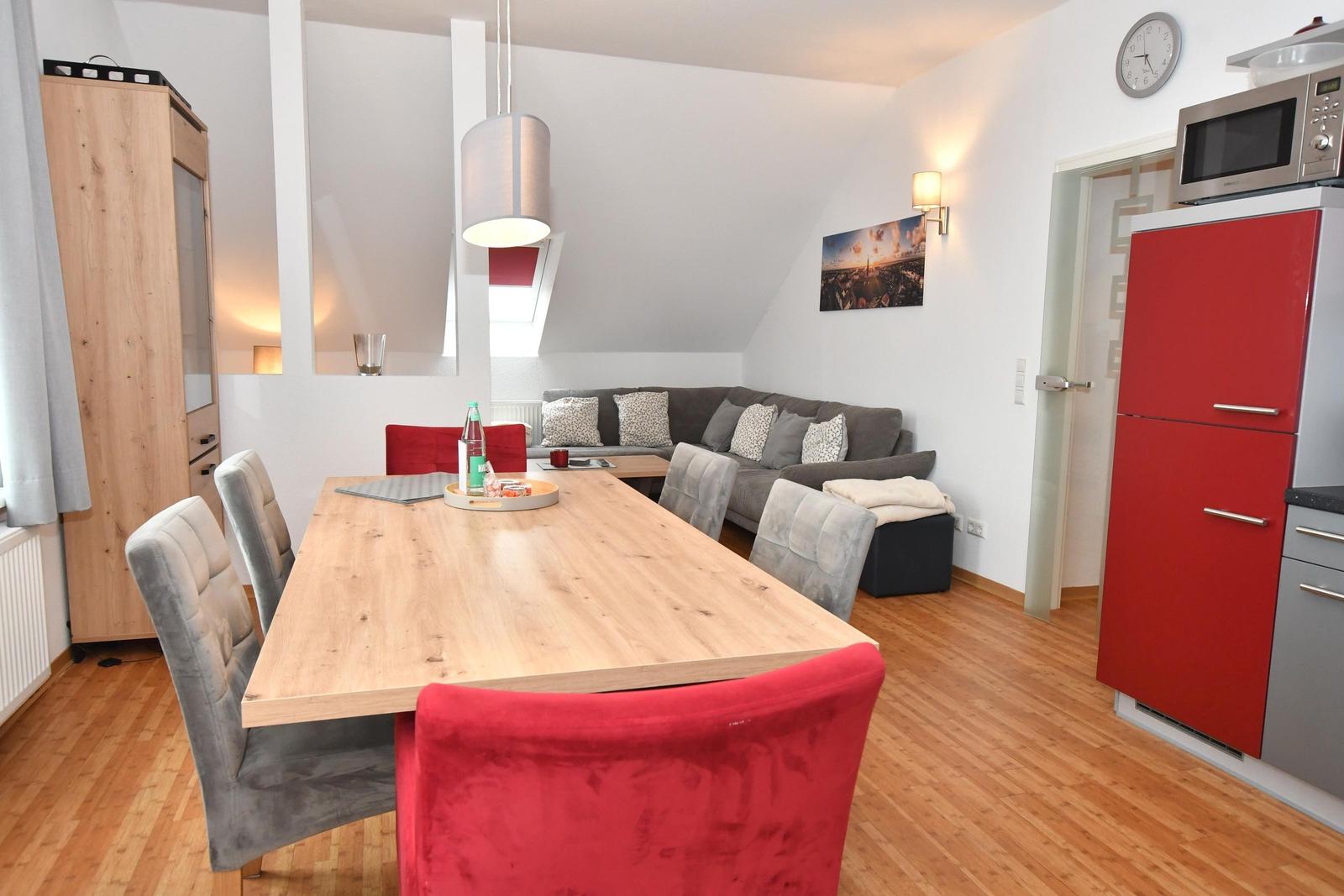 Essbereich mit Holztisch, roten und grauen Stühlen, Sofa und roter Küchenzeile.