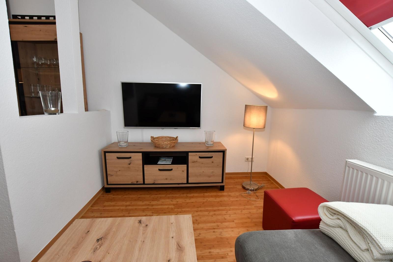 Wohnzimmer mit Fernseher, Holzmöbel und rotem Sitzkissen unter Dachschrägen.
