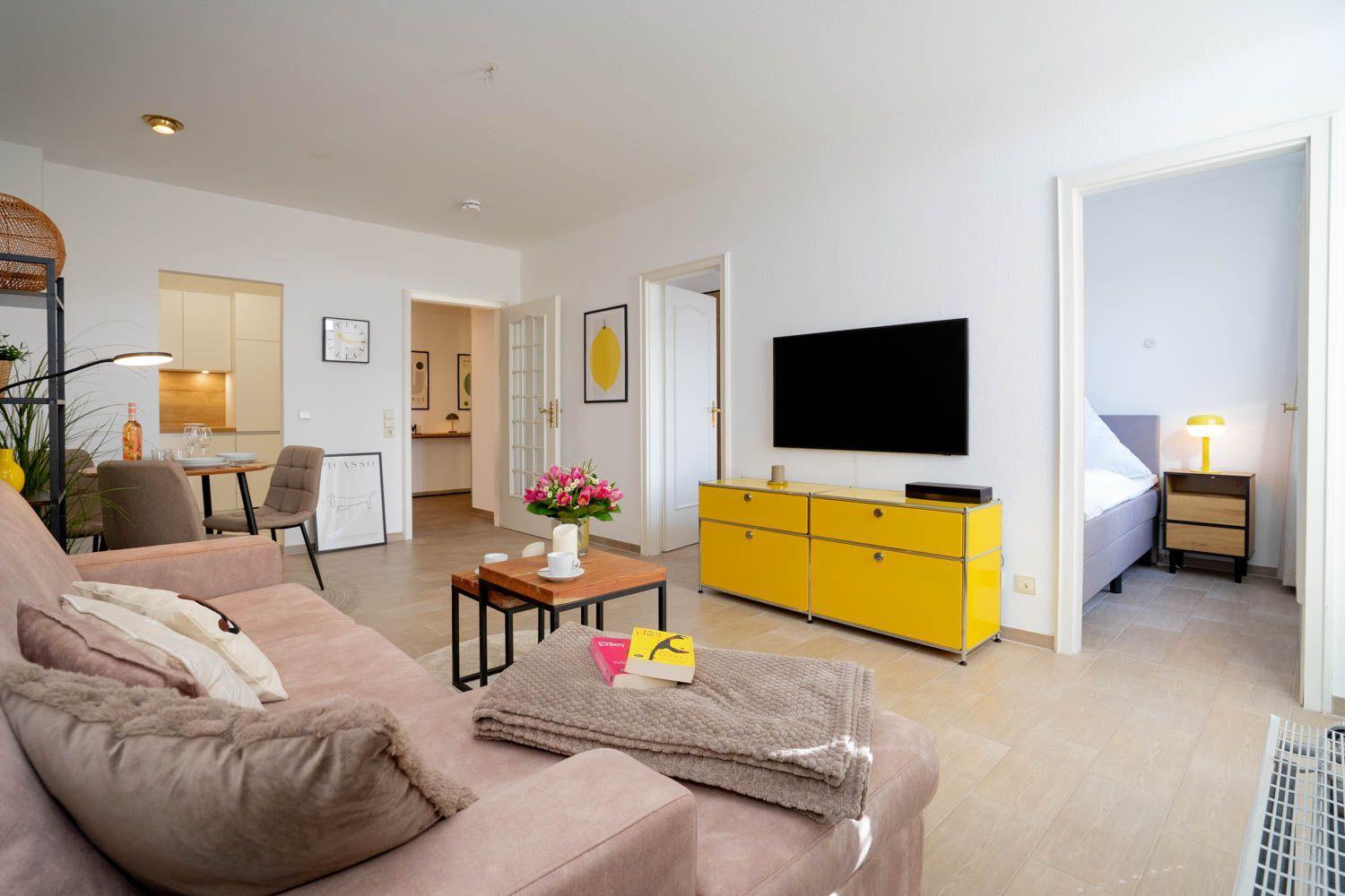 Wohnung mit Sofa, Fernseher und gelbem Schrank. Im Hintergrund ist das Schlafzimmer sichtbar.