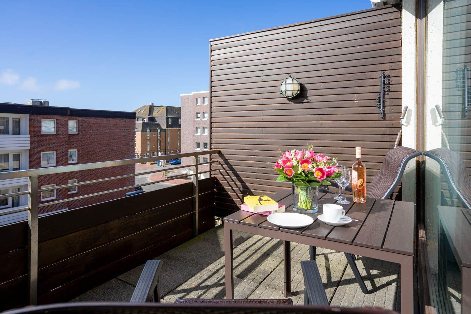 Terrasse mit Tisch, Stühlen, Blumen und Flasche. Blick auf Gebäude und Himmel.