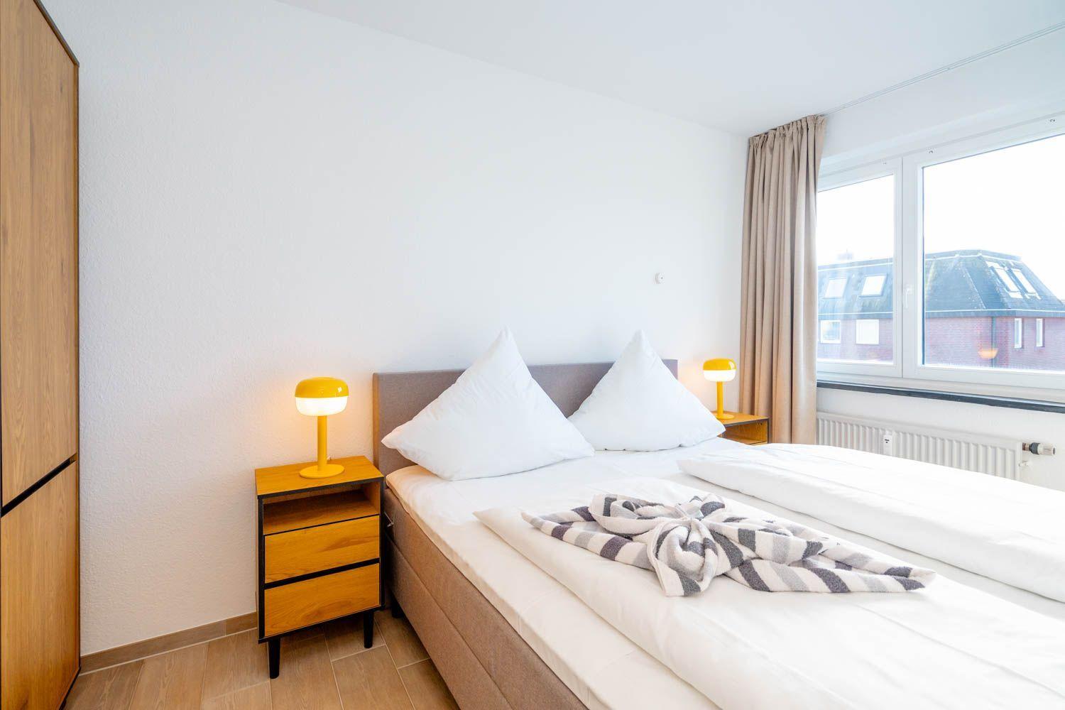 Schlafzimmer mit Bett, Nachttisch und Fenster. Auf dem Bett liegt eine Decke.