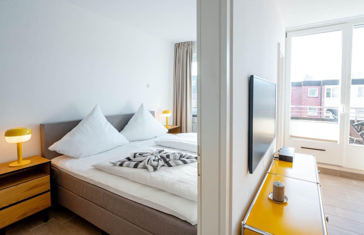 Schlafzimmer mit Bett, Nachttisch und Fernseher. Durchgang zur Terrasse mit Blick auf Gebäude.