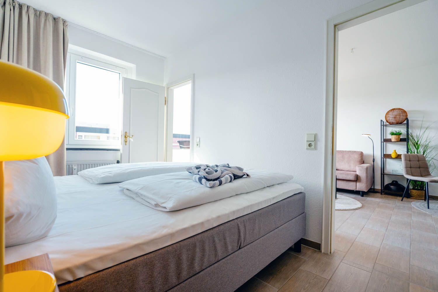 Schlafzimmer mit Bett, Fenster und Tür zum Wohnbereich. Gelbe Lampe und Holzboden.