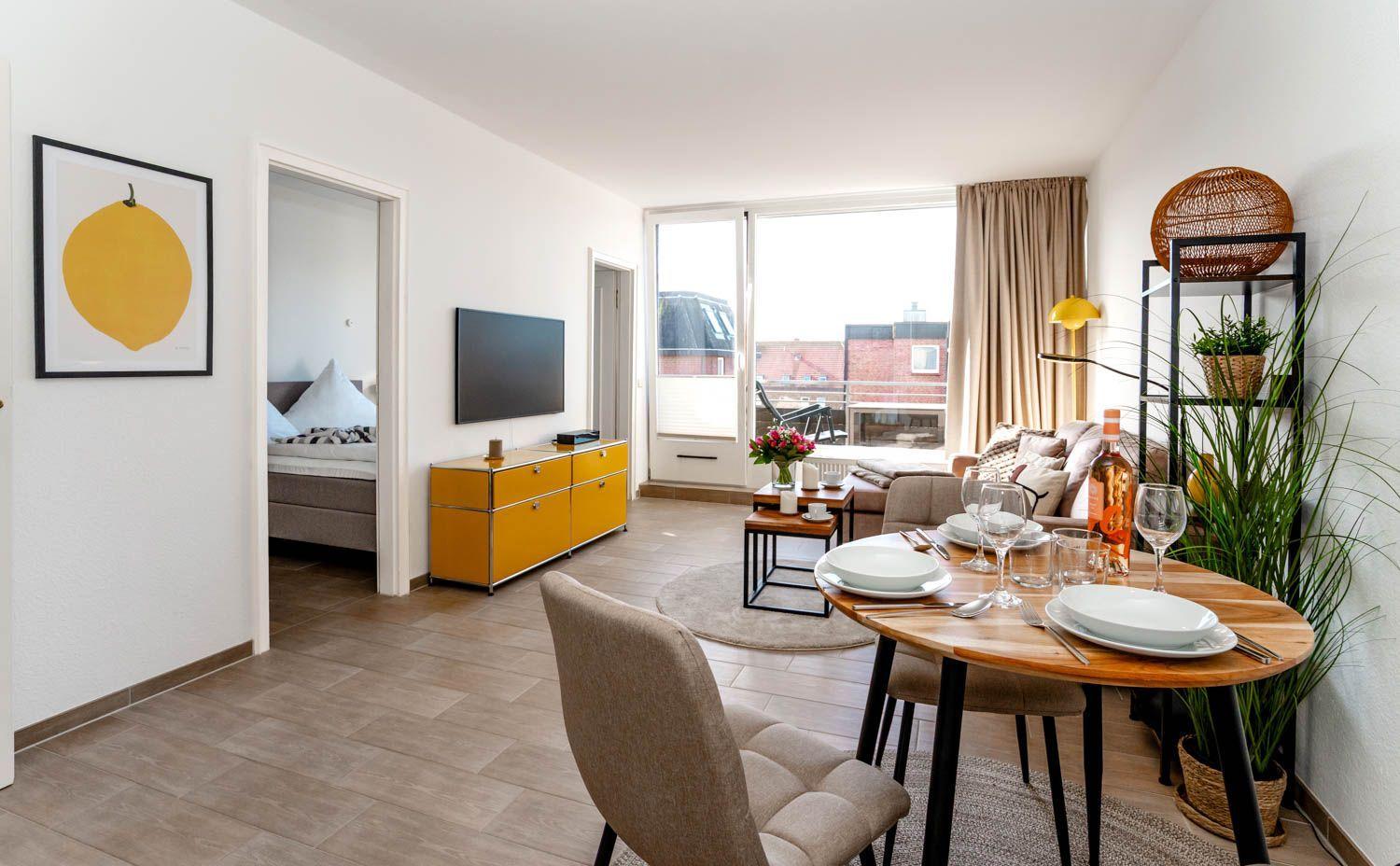 Wohnung mit Essbereich, Schlafzimmer und Balkon. Gelber Schrank, Fernseher und Pflanzen.