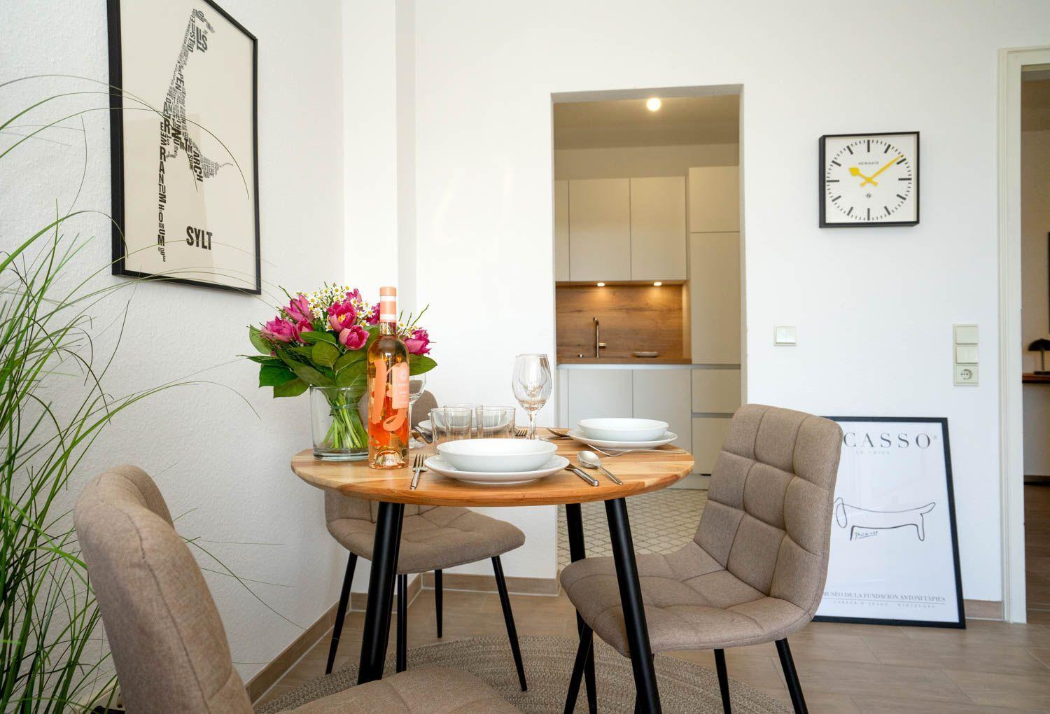 Rundtisch mit Stühlen, Blumen und Flasche. Küche im Hintergrund. Wandbilder und Uhr sichtbar.