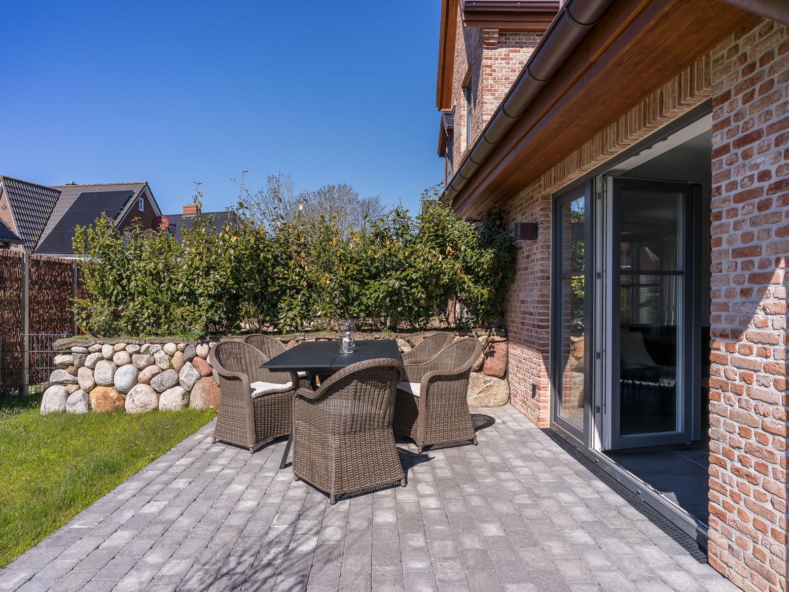 Terrasse mit Tisch und Stühlen neben Haus mit Steinmauer und Hecke.