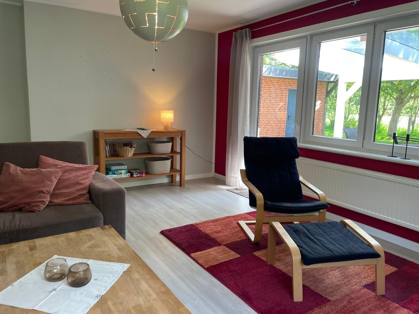 Wohnzimmer mit Sofa, Sessel und großem Fenster. Holzregal und Teppich sind sichtbar.