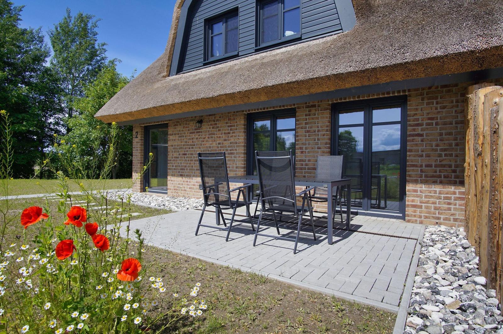Haus mit Strohdach, Terrasse mit Tisch und Stühlen, Blumenbeet mit Mohnblumen.