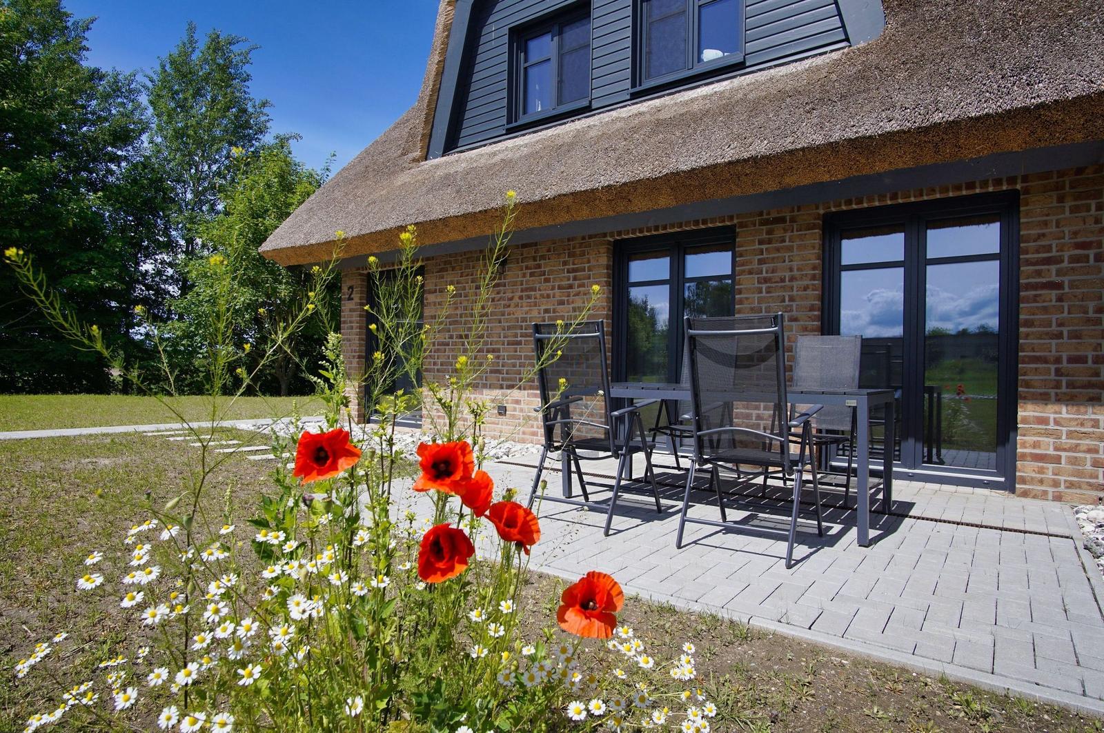 Haus mit Strohdach, Terrasse mit Sitzgruppe und Blumenbeet im Vordergrund.