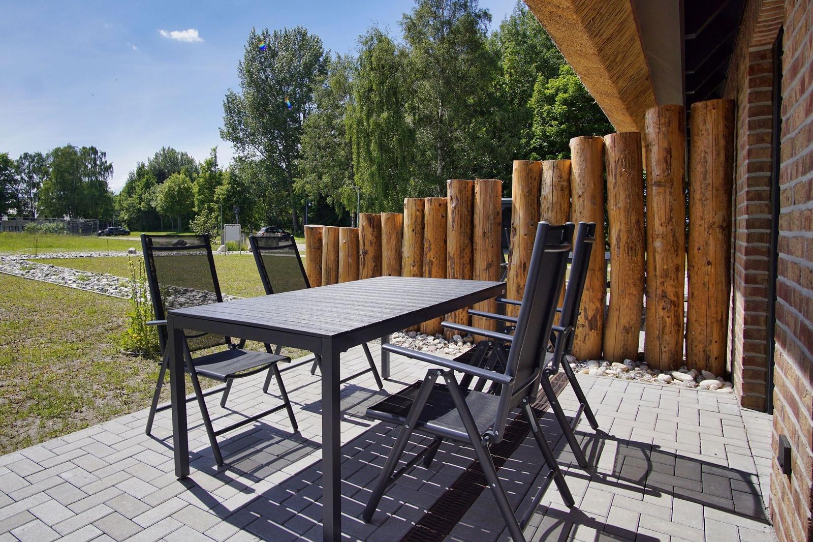 Terrasse mit Tisch und Stühlen, Holzpfosten und Ziegelwand. Grünfläche und Bäume im Hintergrund.
