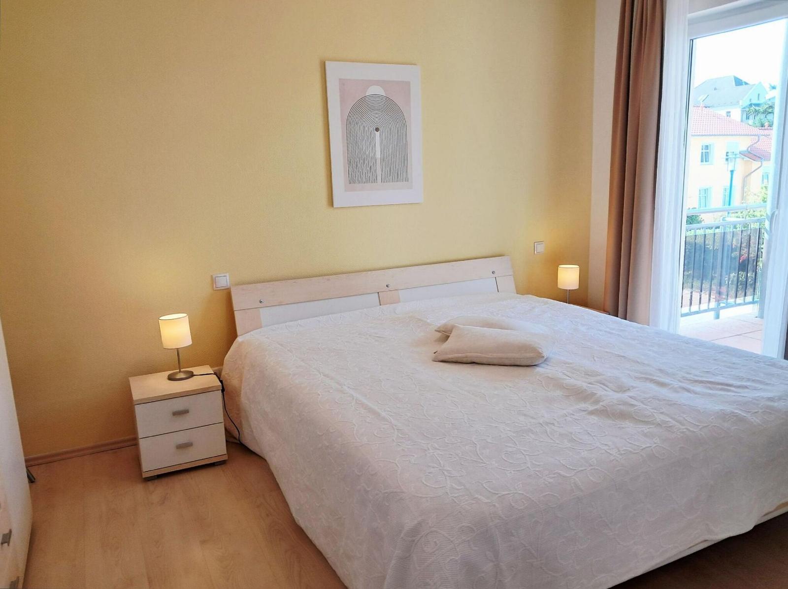 Schlafzimmer mit Bett, Nachttisch und Fenster zum Balkon.