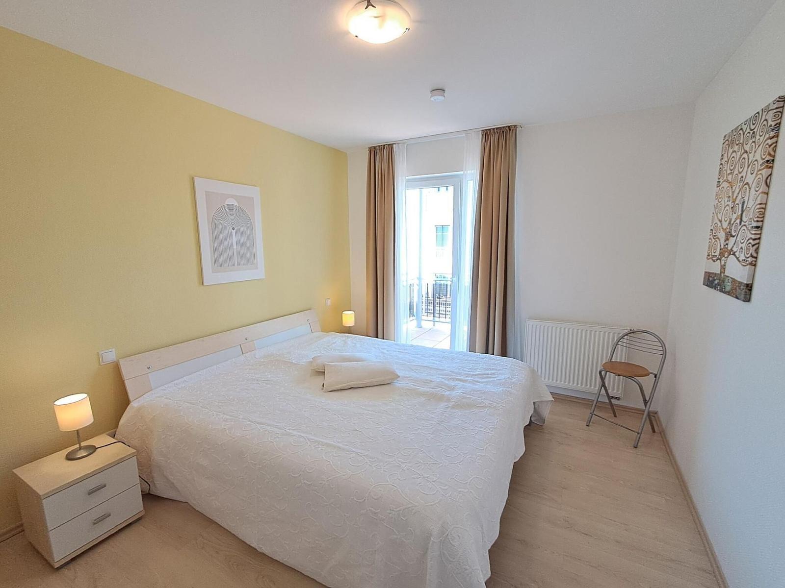 Schlafzimmer mit Bett, Nachttisch, Stuhl und Balkontür.