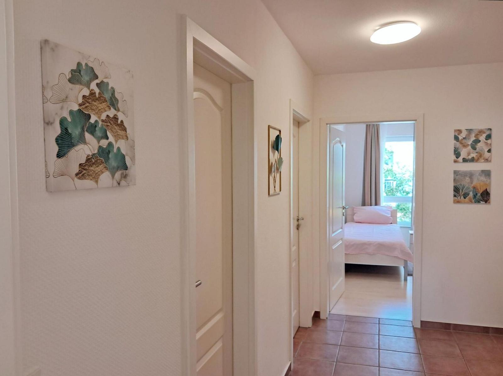 Flur mit Türen und Wandbildern, Blick in Schlafzimmer mit Bett und Fenster.