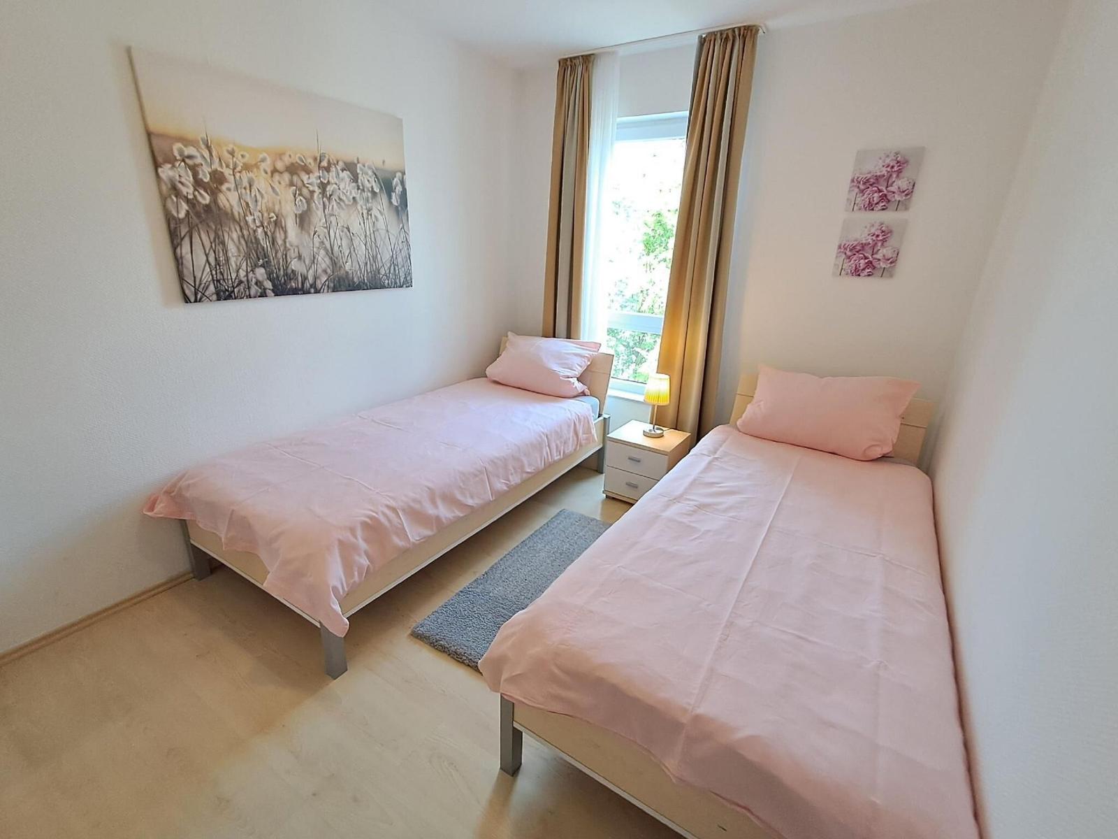 Zwei Einzelbetten mit rosa Bettwäsche, Fenster mit Vorhängen und Wandbilder.