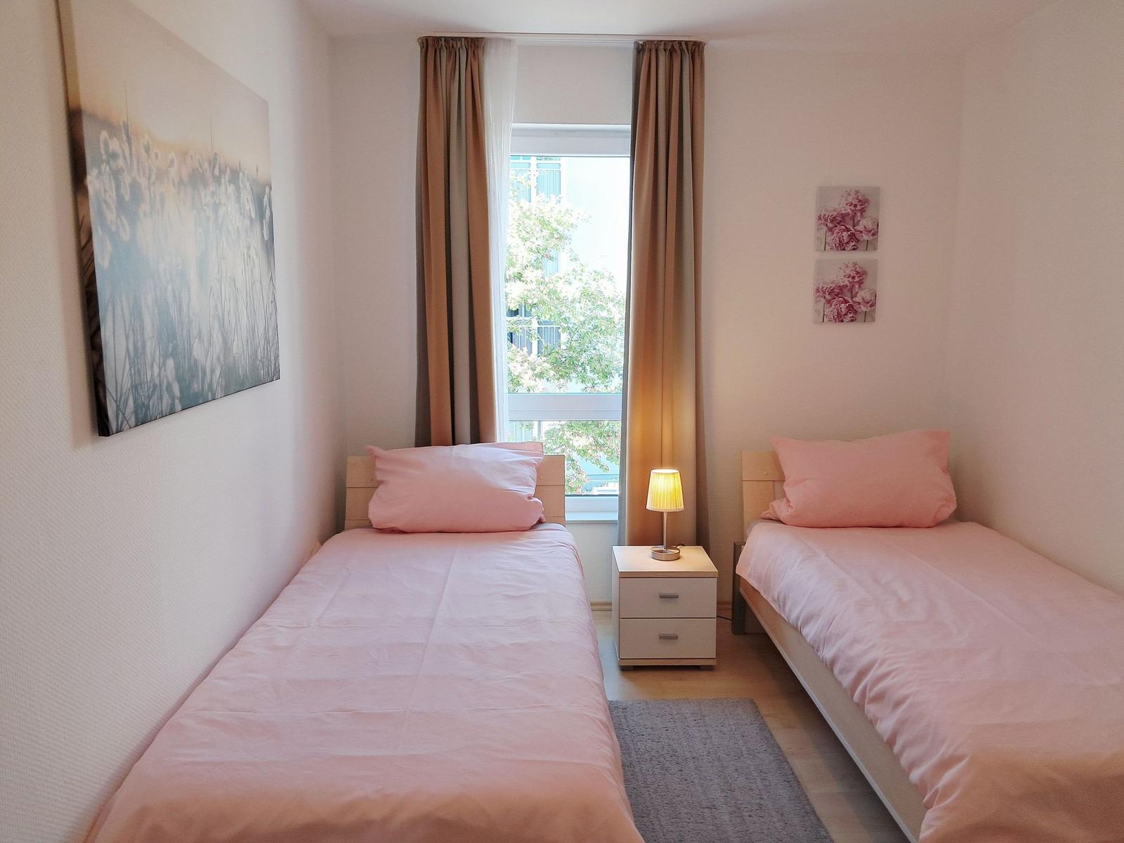 Zwei Einzelbetten mit rosa Bettwäsche, Nachttisch mit Lampe und Fenster mit Blick.