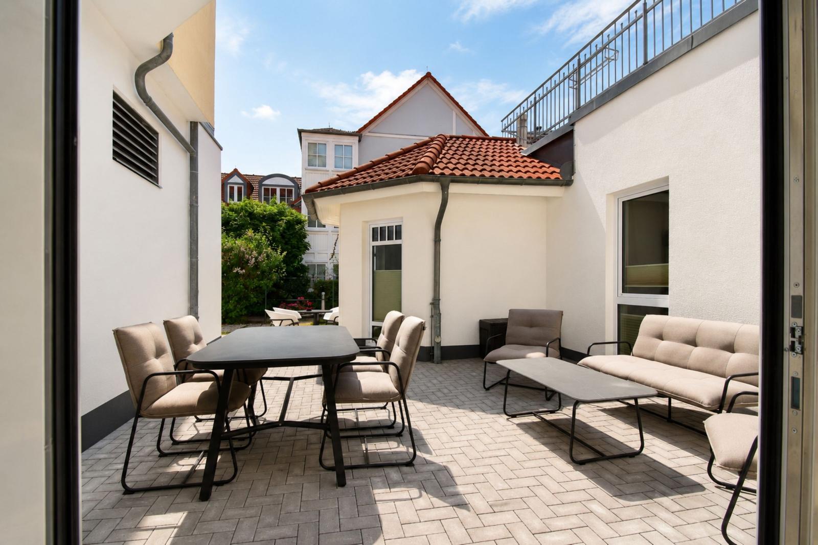 Terrasse mit Sitzgruppe, Tisch und Stühlen. Hintergrund: Haus mit rotem Dach.