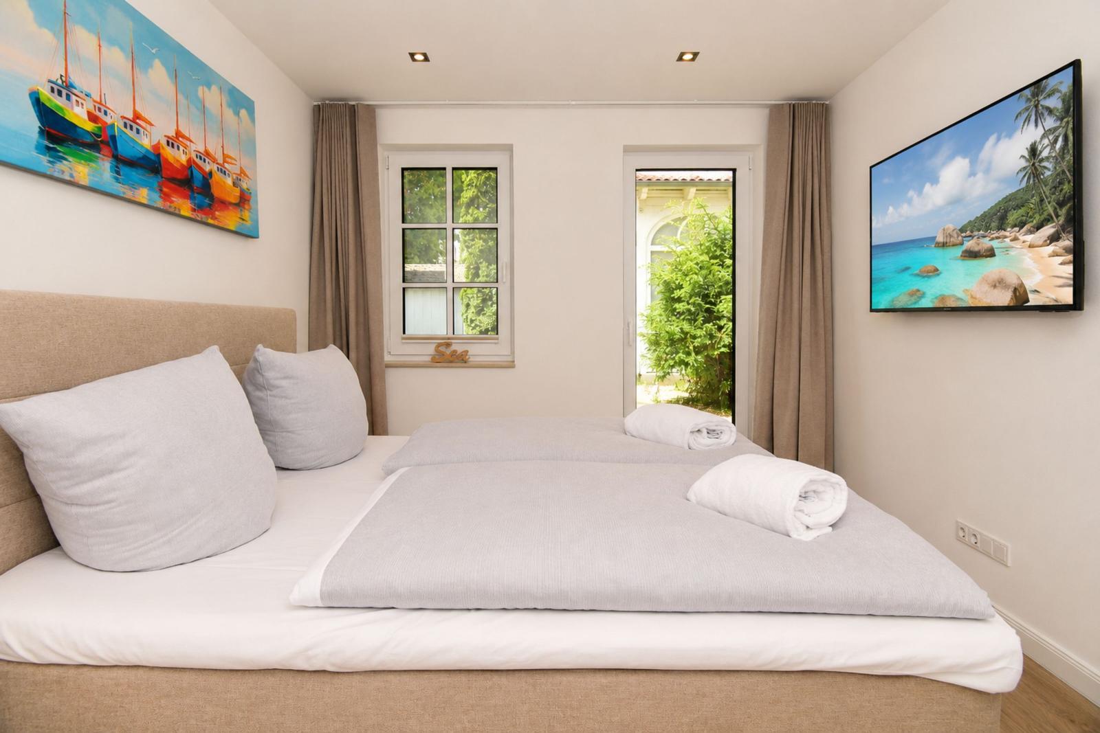 Schlafzimmer mit Bett, Fenster und TV. Wandbilder zeigen Segelboote und Strand.