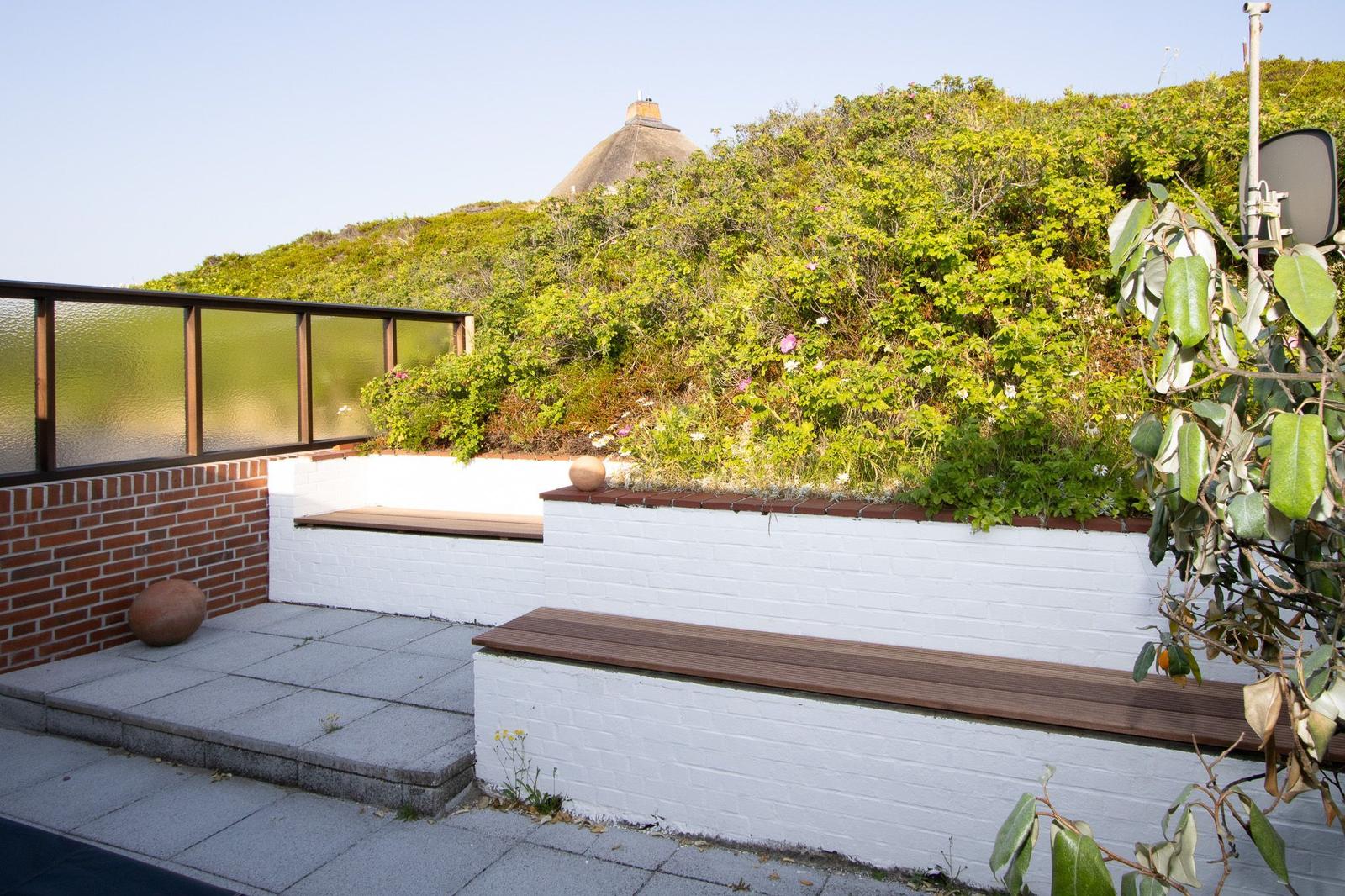 Terrasse mit Sitzbänken, weißen Mauern und grüner Vegetation. Im Hintergrund ist ein Dach zu sehen.