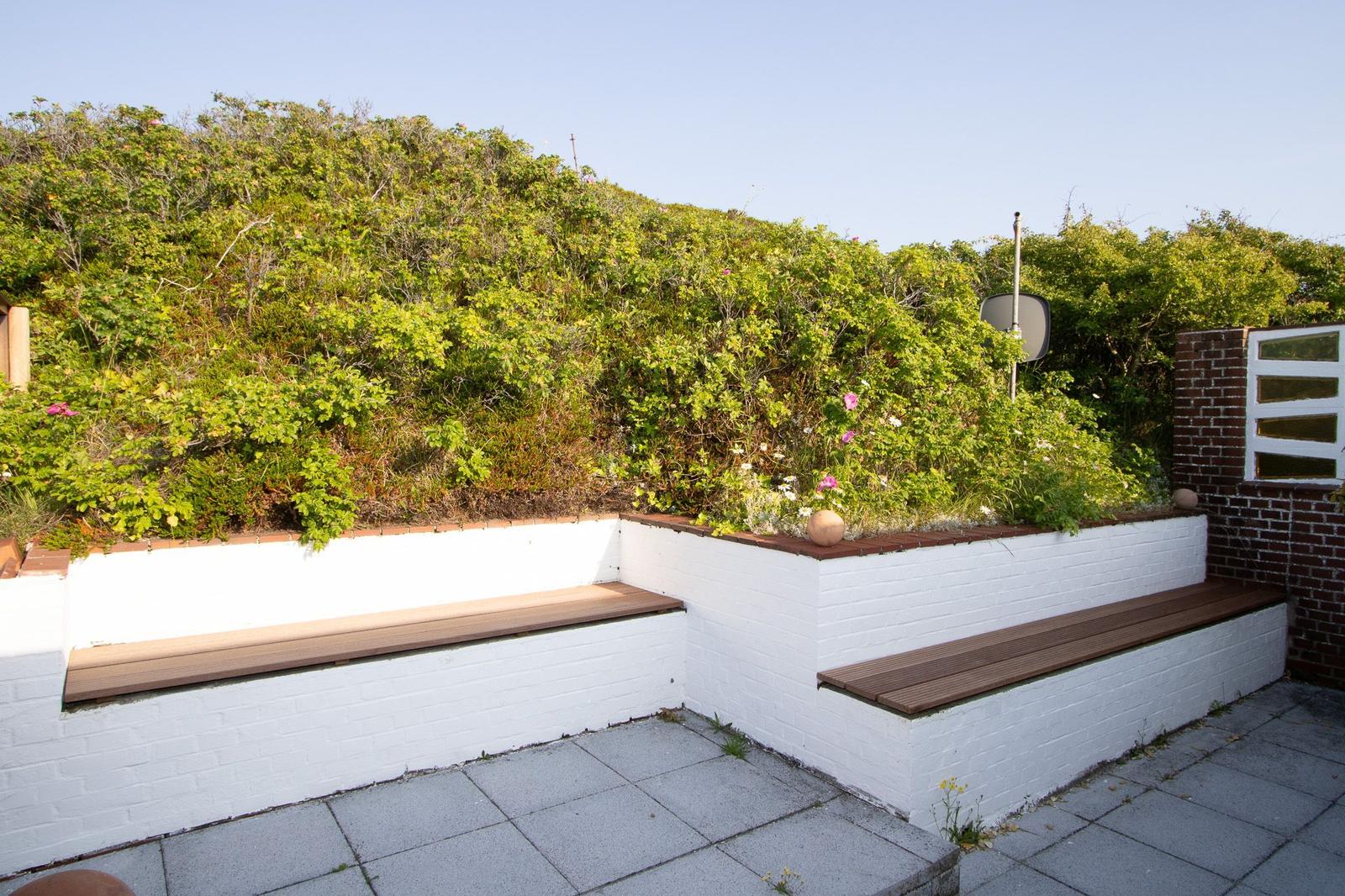 Terrasse mit weißen Mauern und Holzbänken, umgeben von grüner Vegetation und einem Steinweg.