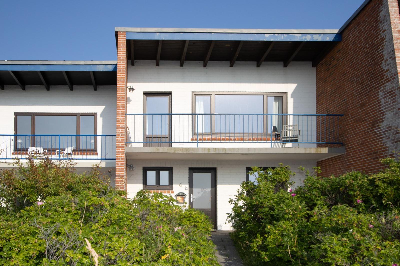 Zweistöckiges Haus mit Balkon, weißen Wänden und roten Ziegelsteinen. Grünflächen vorne.