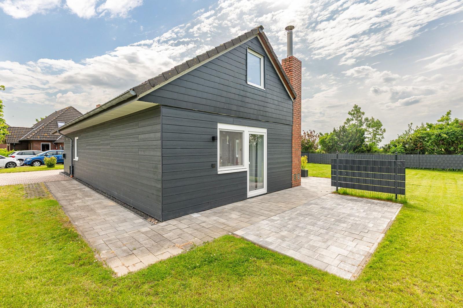 Moderne Villa mit grauer Fassade, Terrasse und Garten. Holzfenster und Ziegelkamin sichtbar.