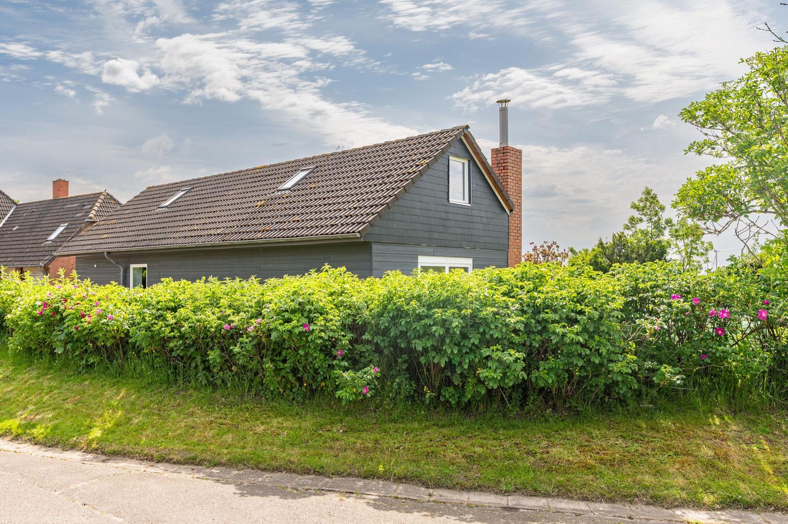 Ein Haus mit grauer Fassade und rotem Schornstein hinter grünen Sträuchern mit rosa Blüten.