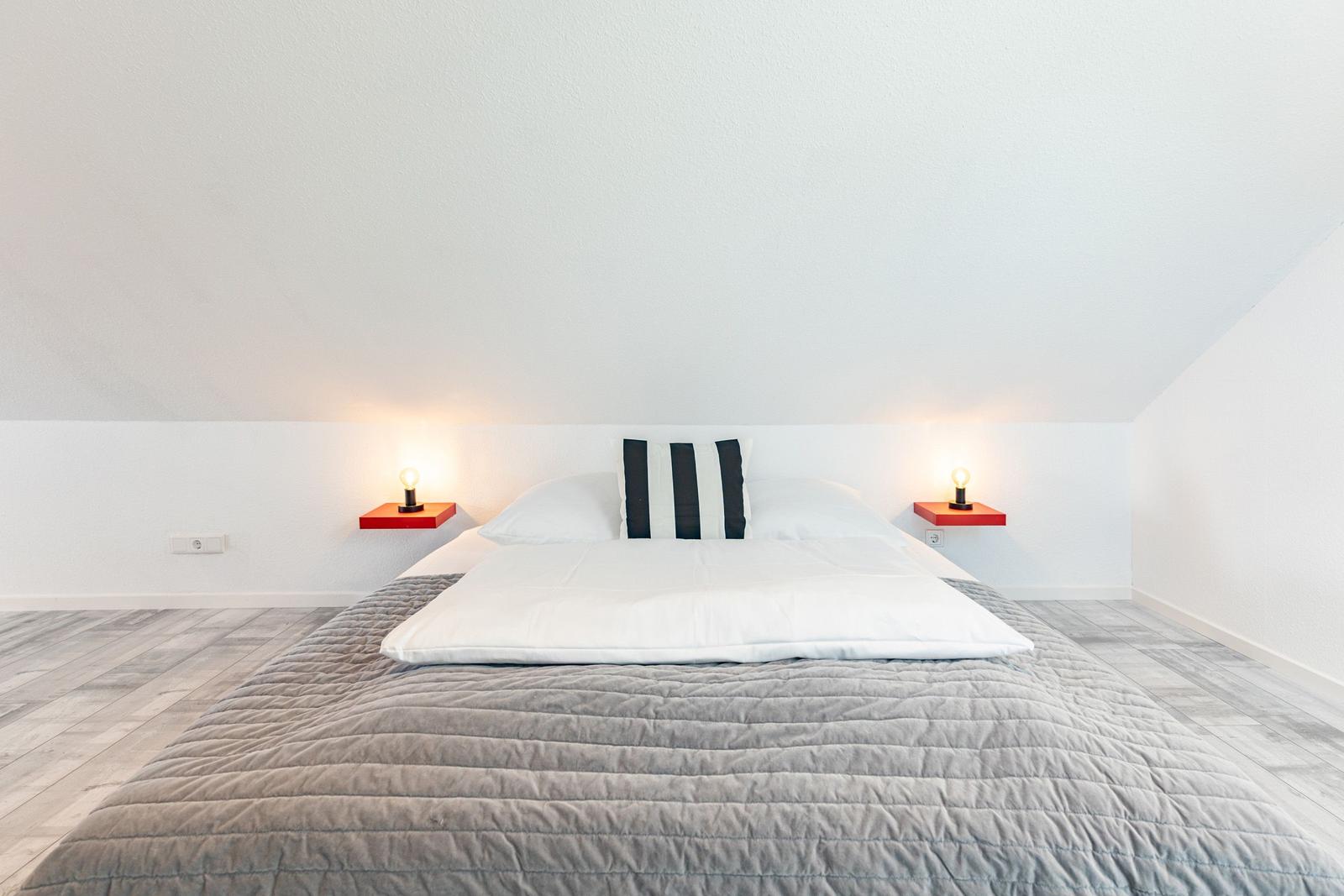 Bedroom with double bed, gray quilt, and two red nightstands with lamps.