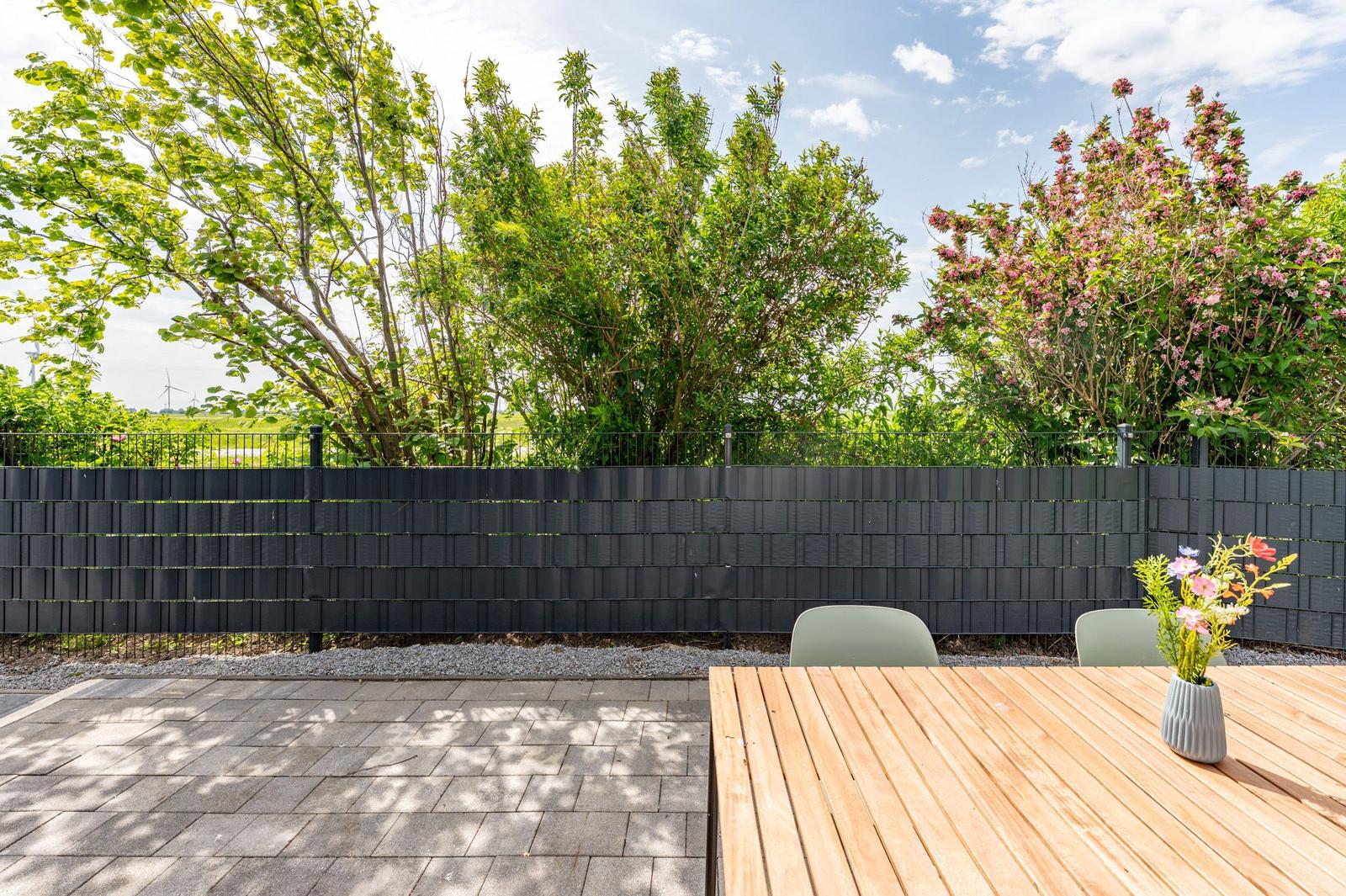 Backyard with table, chairs, and flowers. Black wall, trees, and flowers.