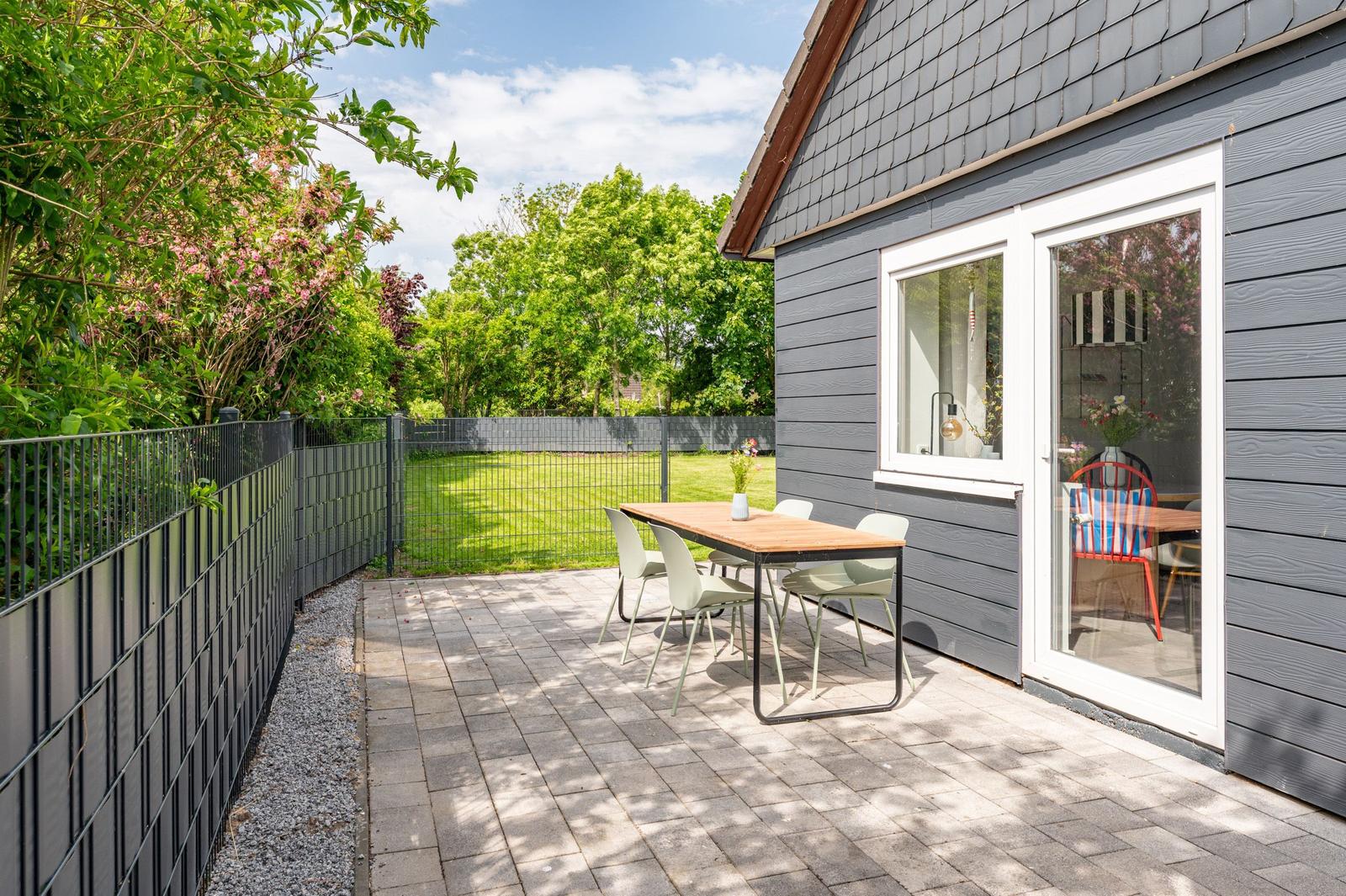 Terrace with table and chairs beside gray house. Backyard with grass and fence.