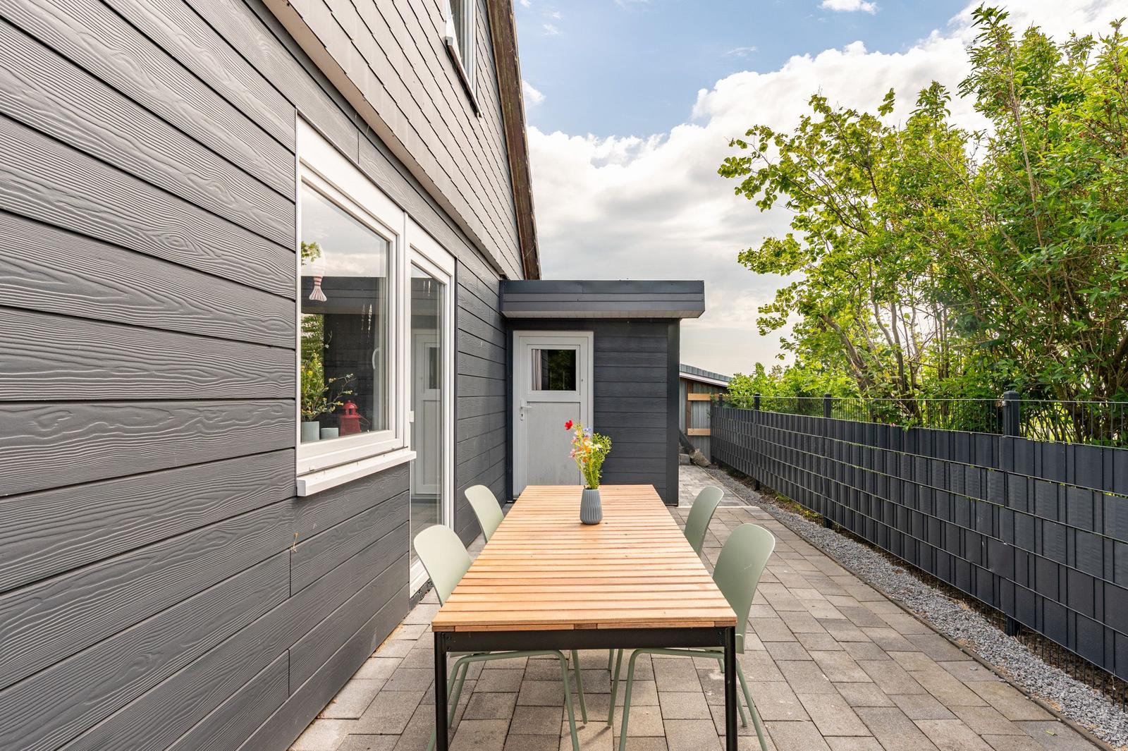 Outdoor area with table and chairs in front of gray wooden house. Greenery and fence in background.